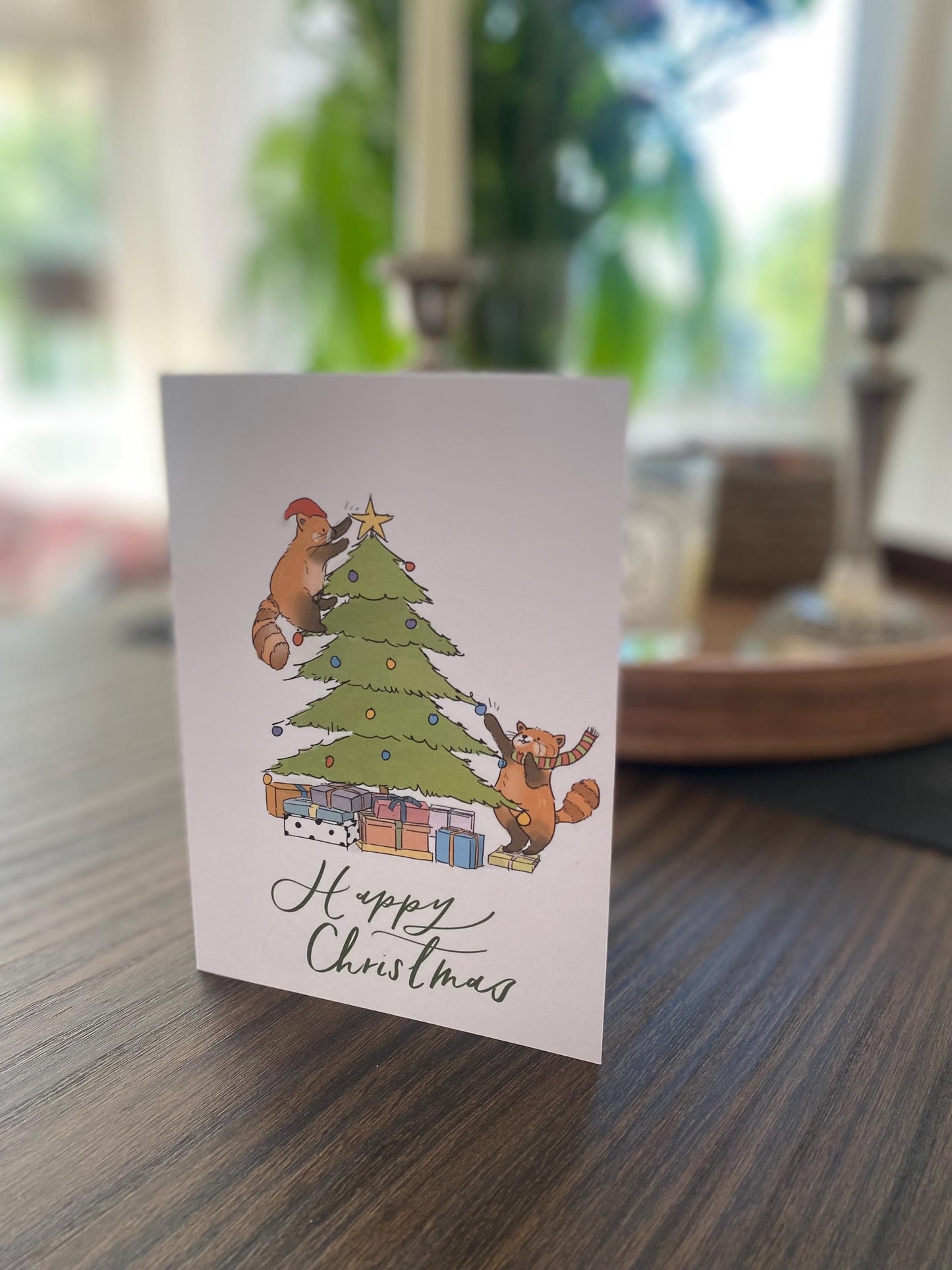 Christmas card with cartoon animals decorating a tree on a wooden table.