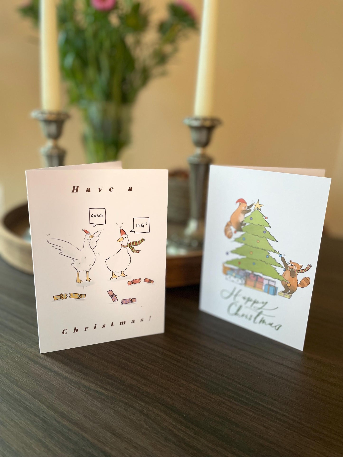 Two Christmas cards on a table with candles and flowers in the background