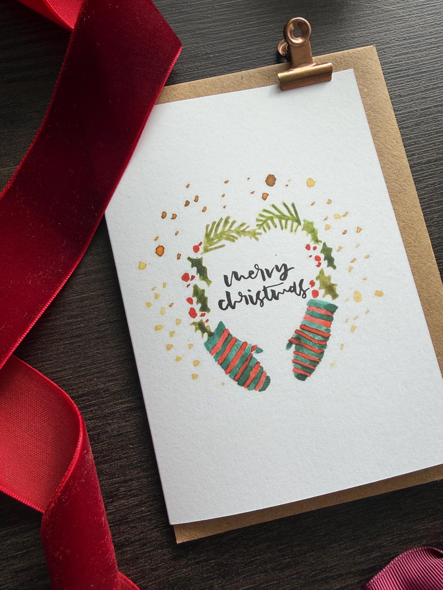 Christmas card with stockings and wreath design, tied with a red ribbon on a wooden surface.