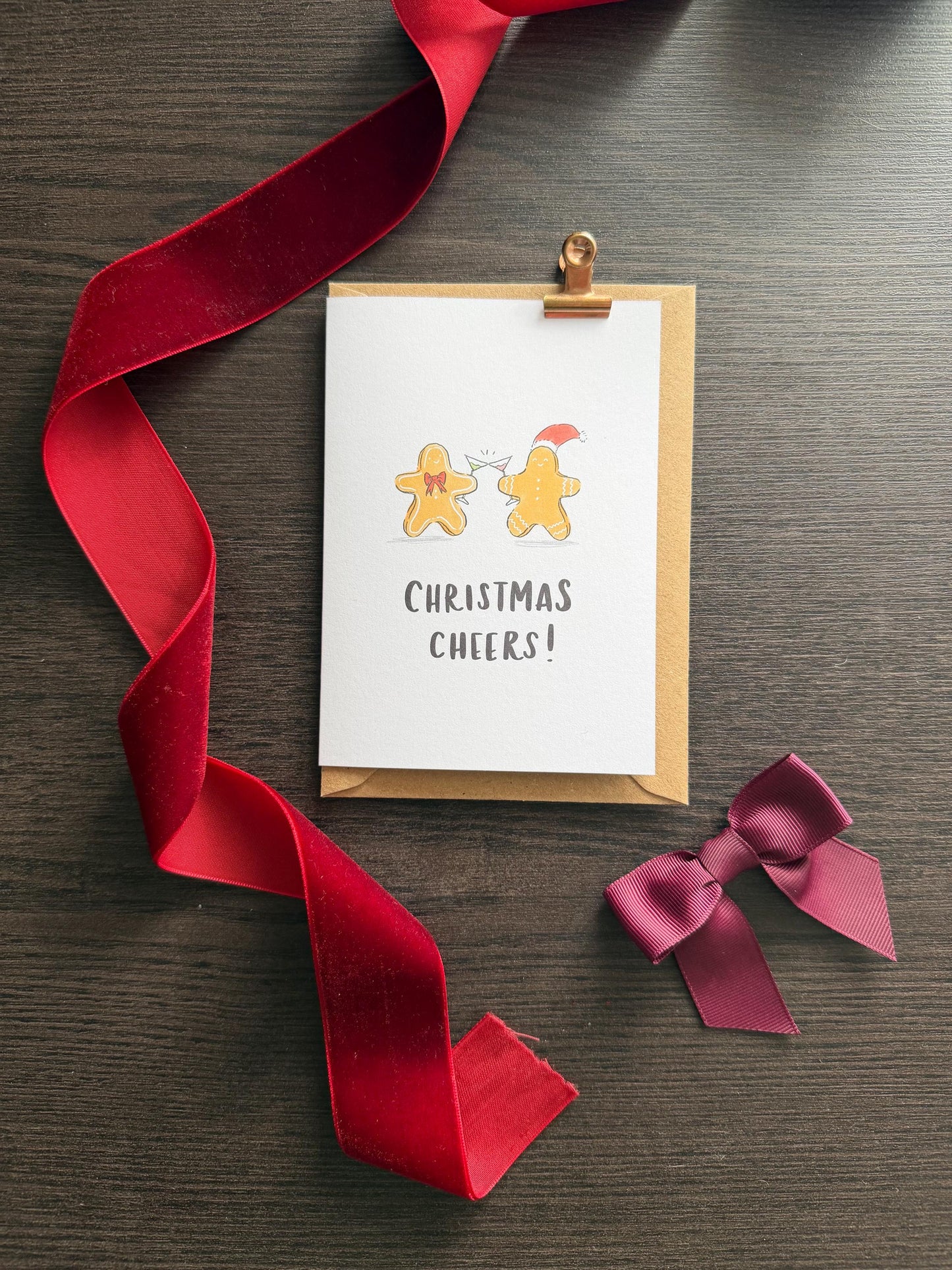 Greeting card with 'Christmas Cheers!' text and gingerbread men illustration, attached to a red ribbon on a wooden surface.