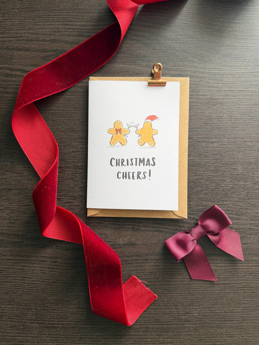 Greeting card with 'Christmas Cheers!' text and gingerbread men illustration, attached to a red ribbon on a wooden surface.
