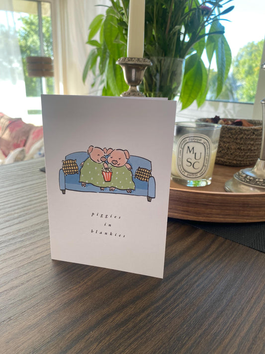 Card with cartoon pigs on a couch on a wooden table with a blurred background