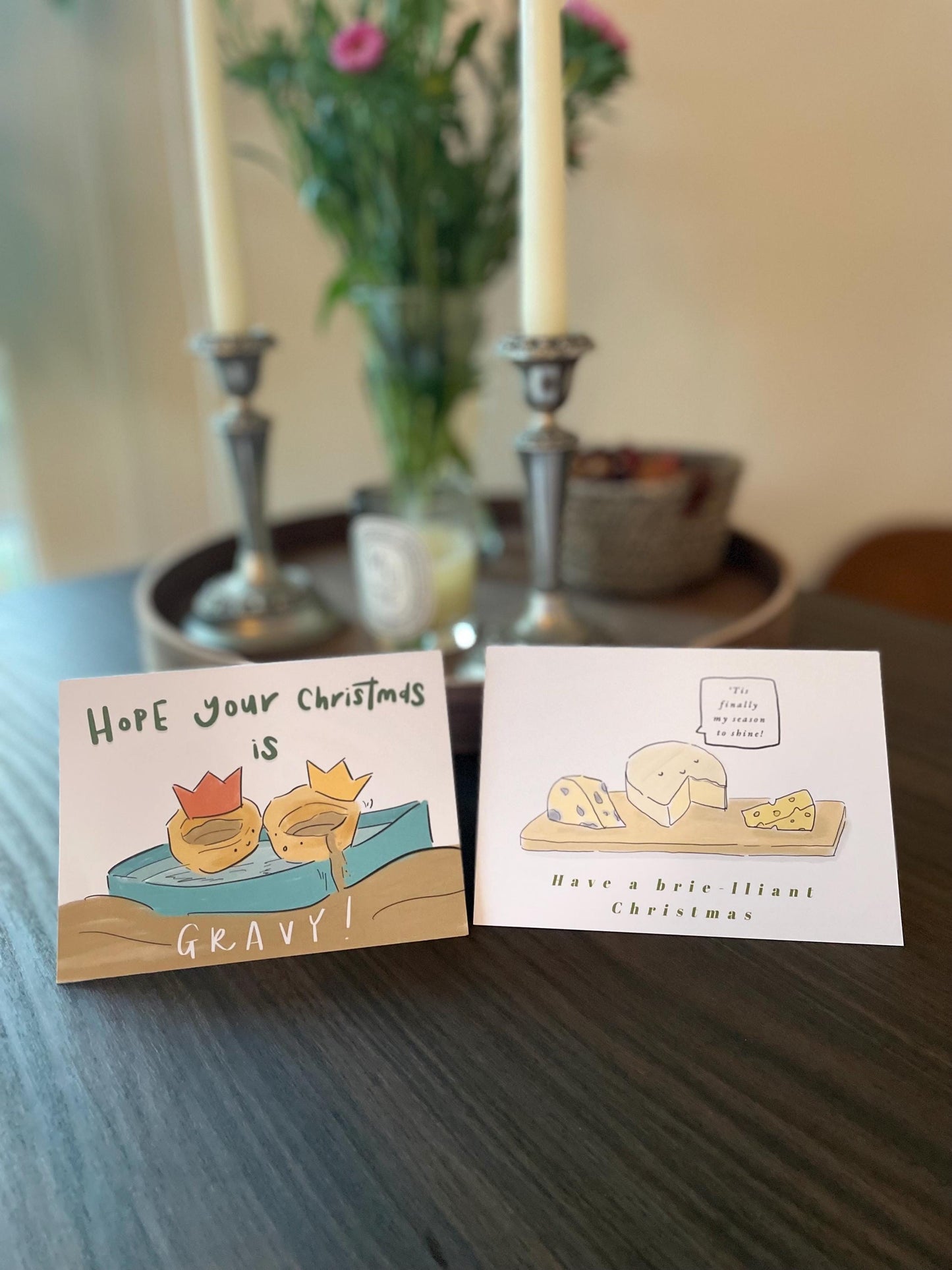 Two Christmas-themed cards on a table with candles and flowers in the background.