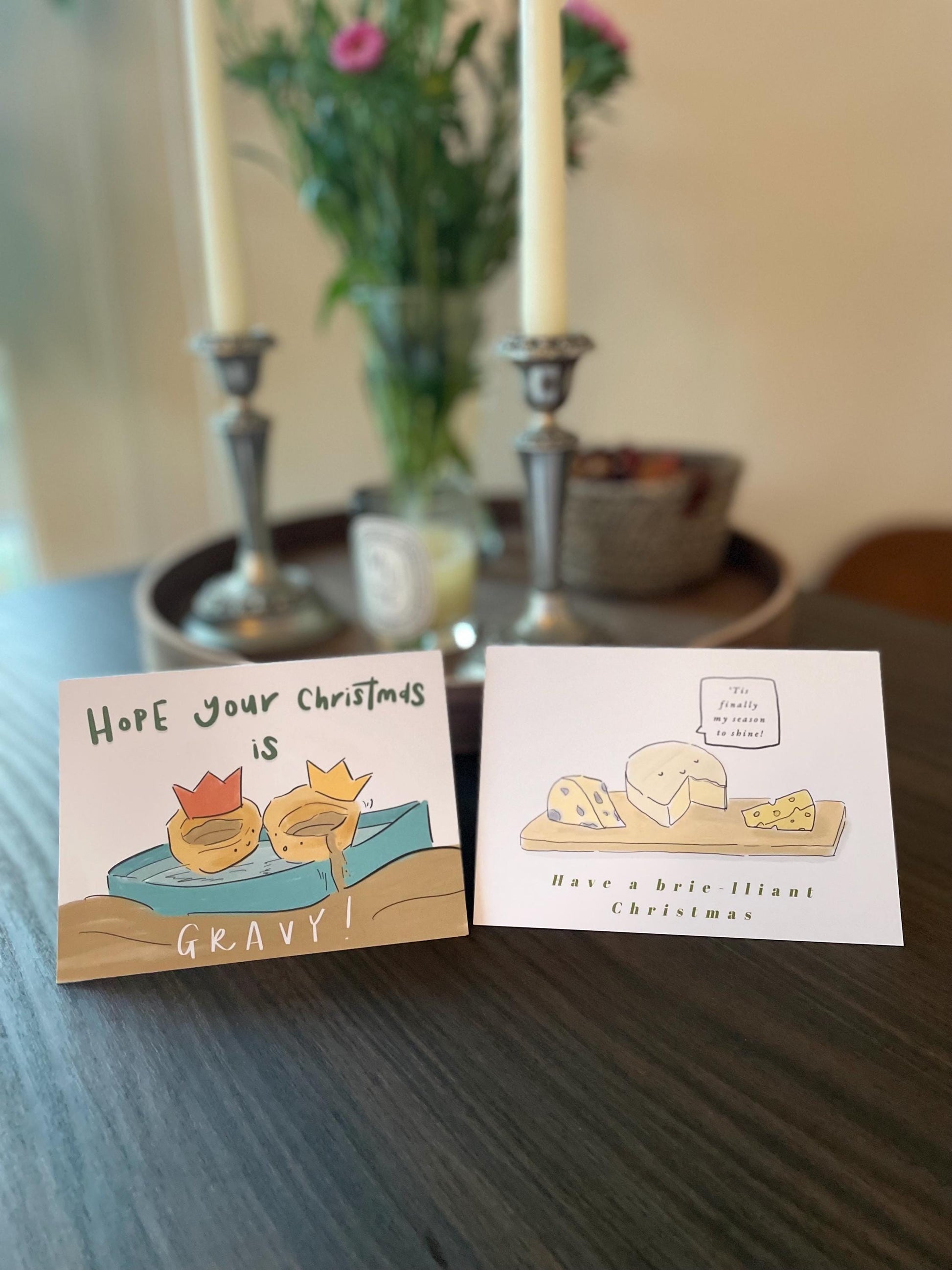 Two Christmas-themed cards on a table with candles and flowers in the background.