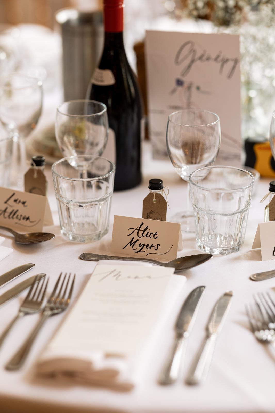 Table setting with wine glasses, bottles, and place cards on a white tablecloth.