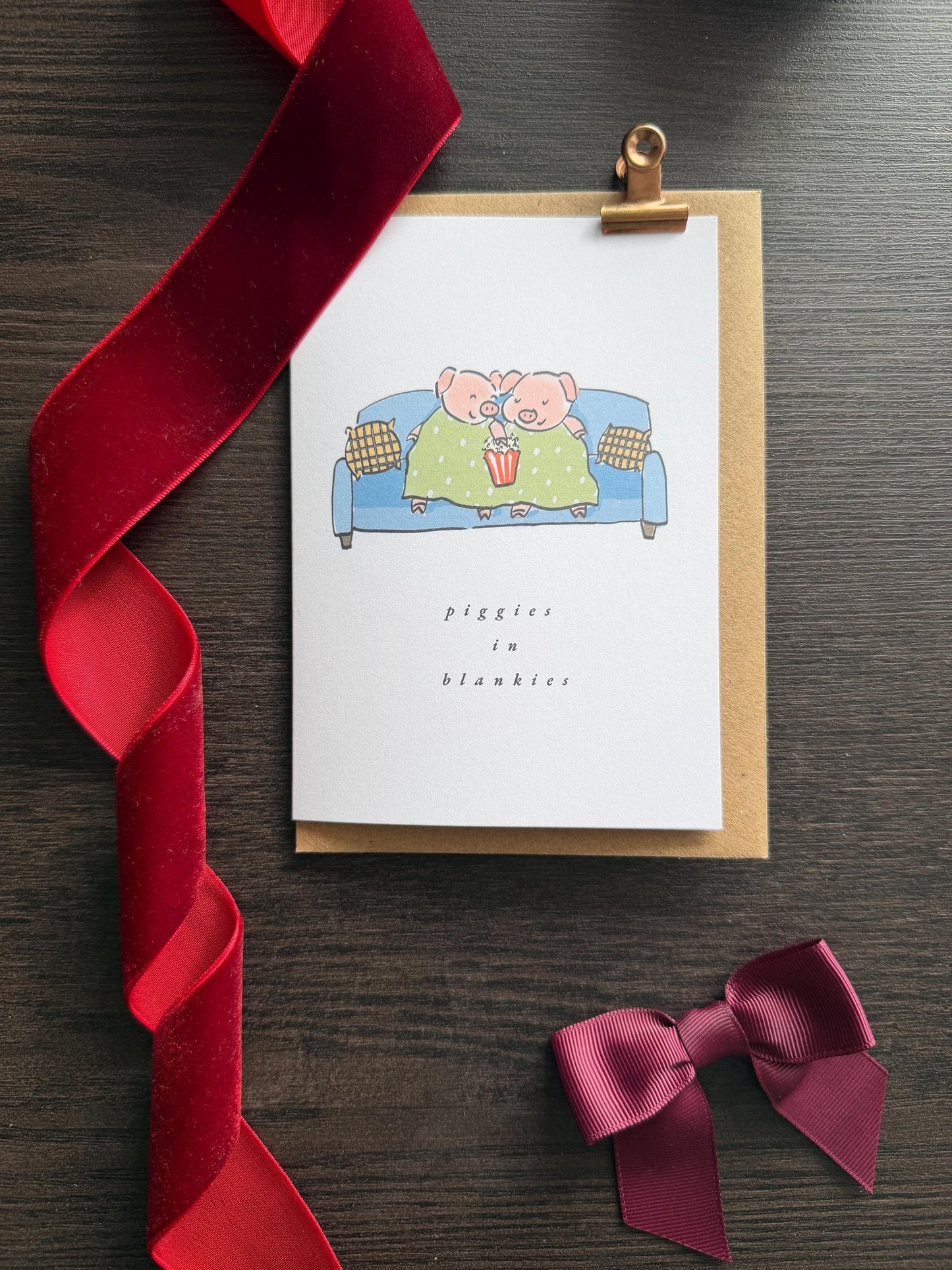 Greeting card with cartoon pigs on a couch, red ribbon, and purple bow on a wooden surface