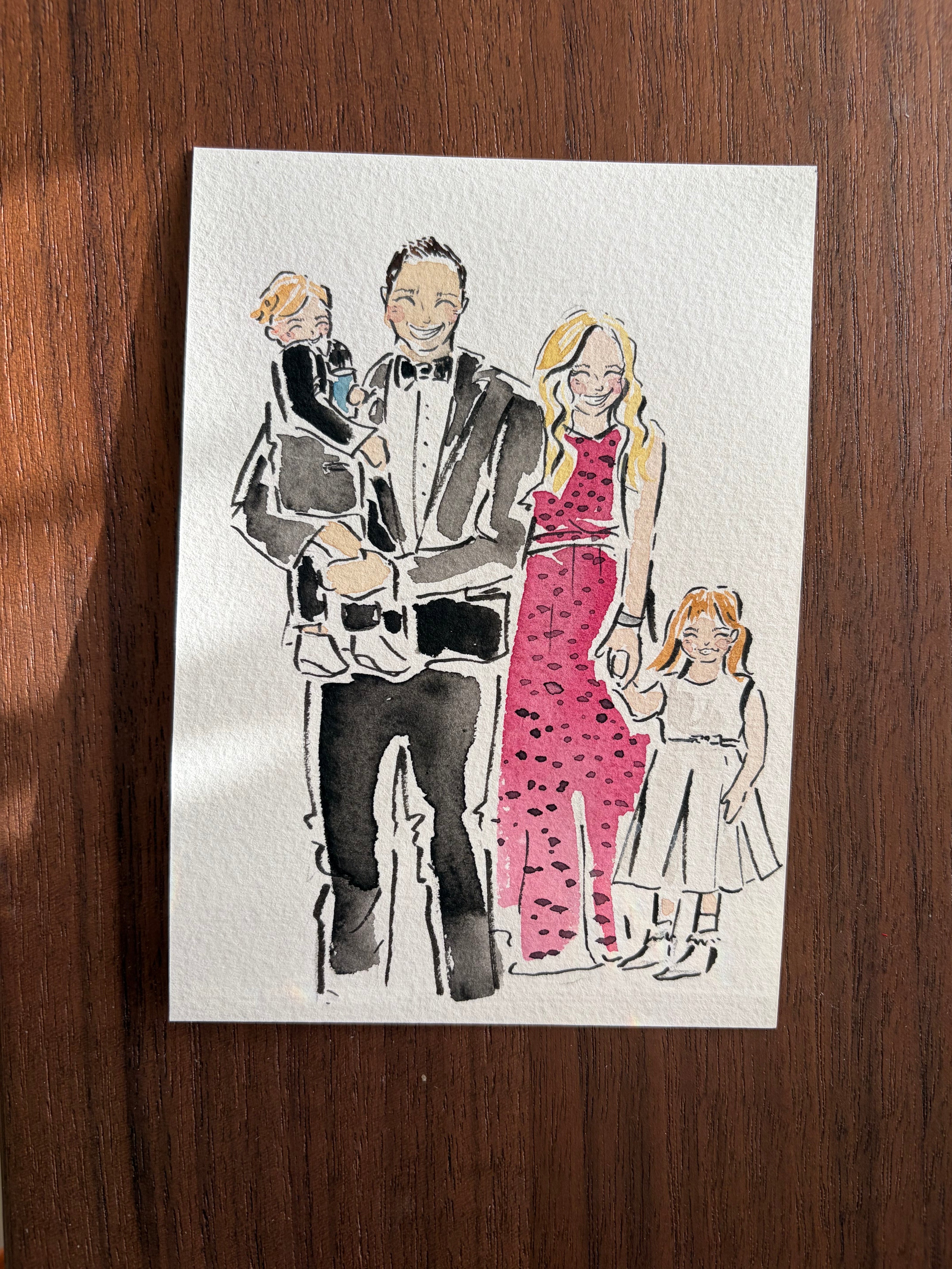 Illustration of a family on a white card with black, pink, and gray details, placed on a wooden surface.
