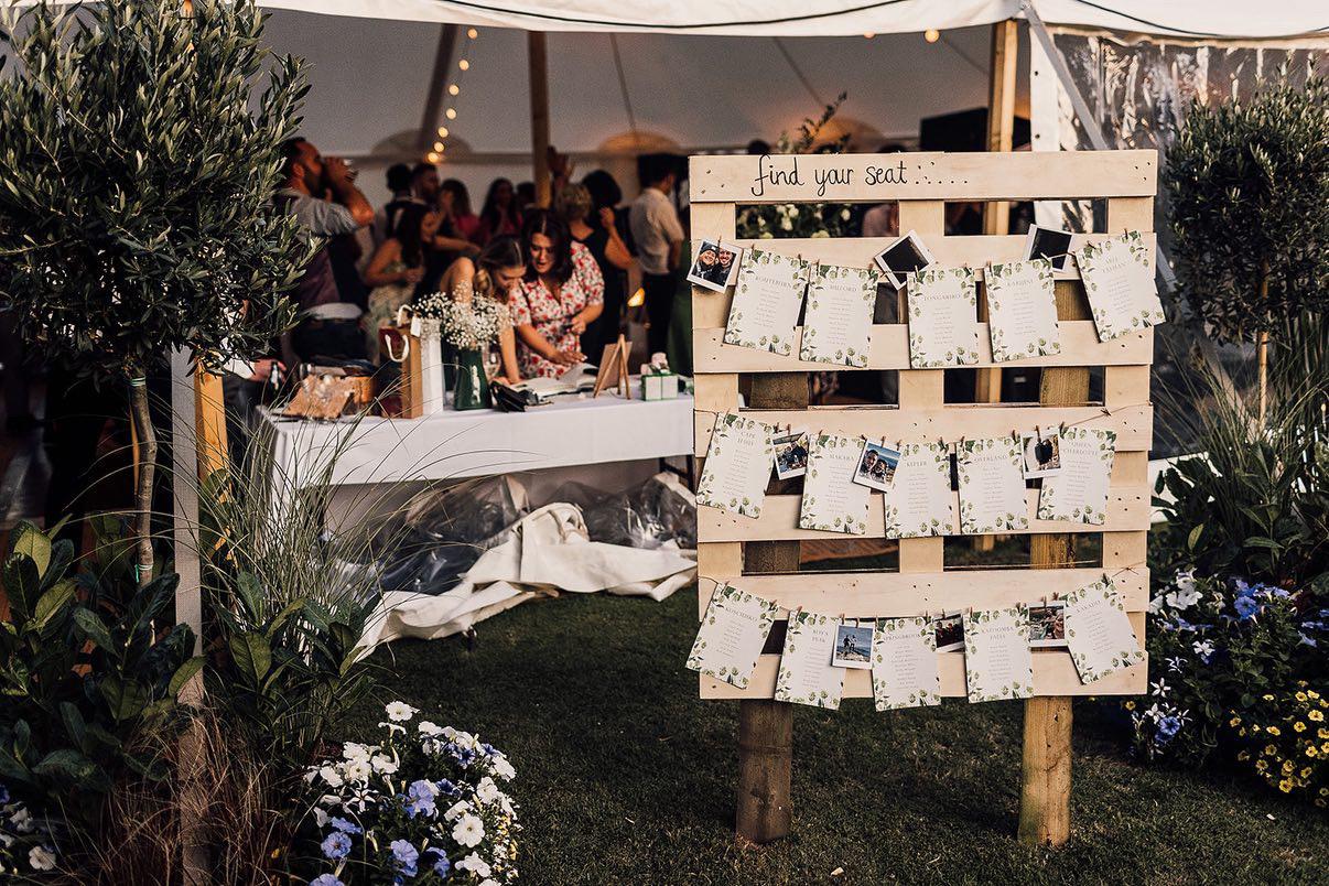 Wooden seating chart at an outdoor event with people in the background.