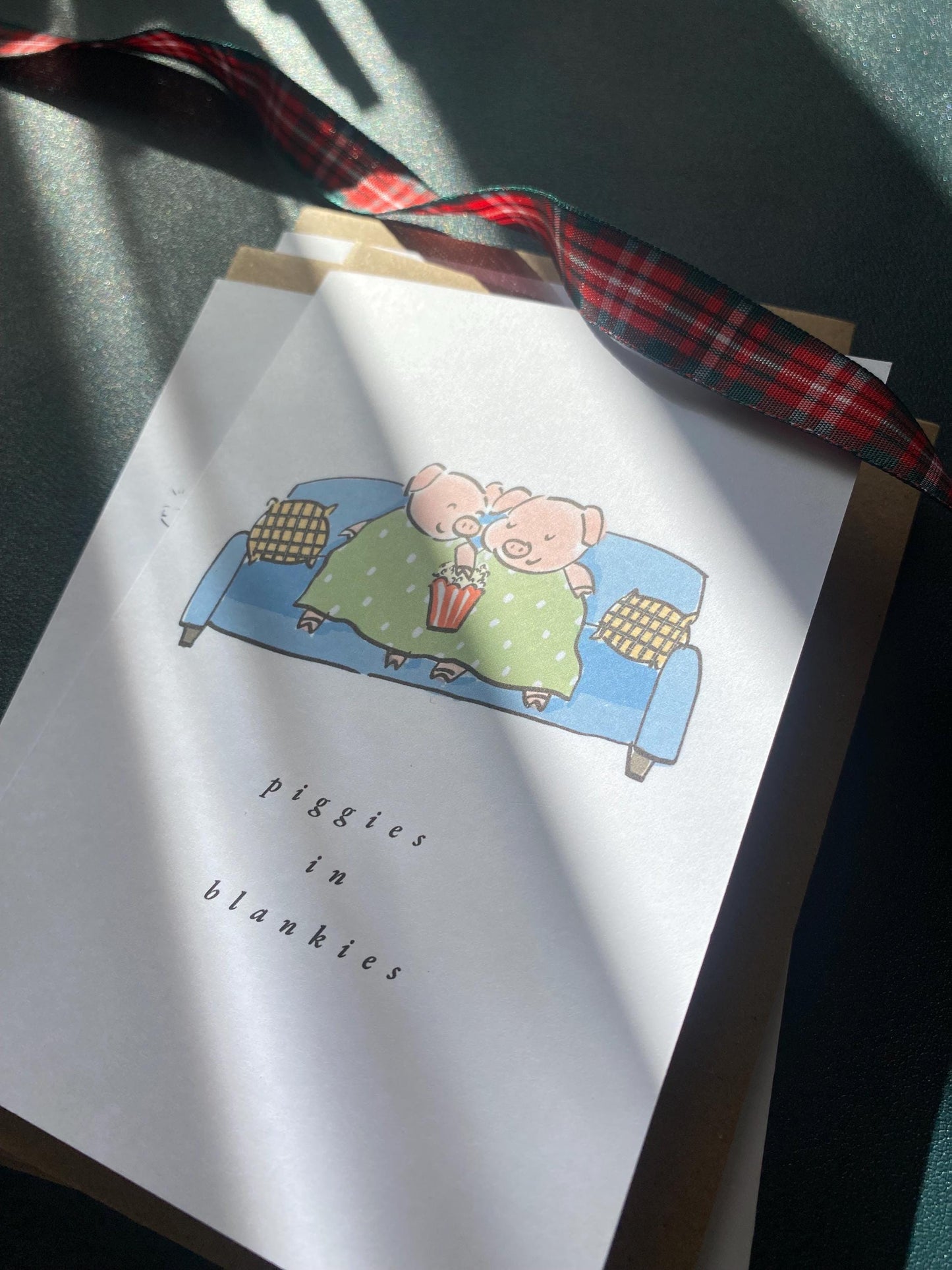 Card with a cartoon pig on a couch and plaid ribbon, titled 'Piggies in Blankies'.